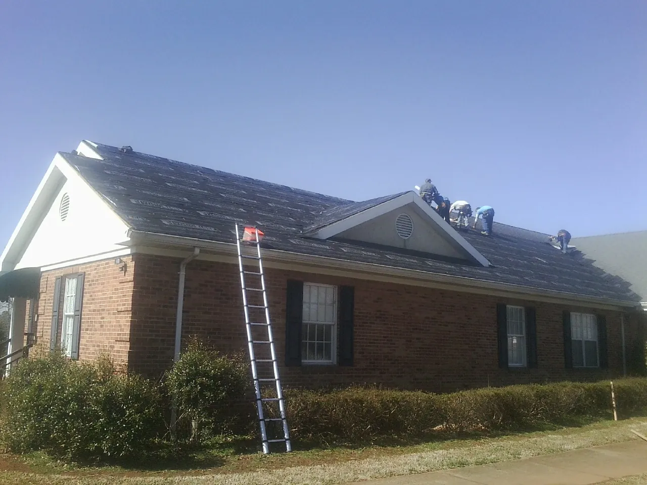 Crew tearing shingles off a brick ranch roof with a ladder set — Emergency Roof Repair work by our crew in Lunenburg